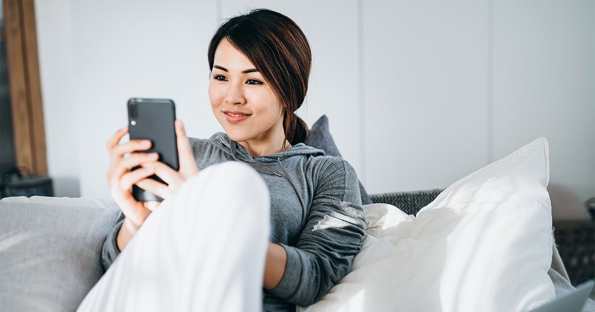 Sorridente giovane donna asiatica che utilizza lo smartphone mentre giace a letto, con il computer portatile a lato